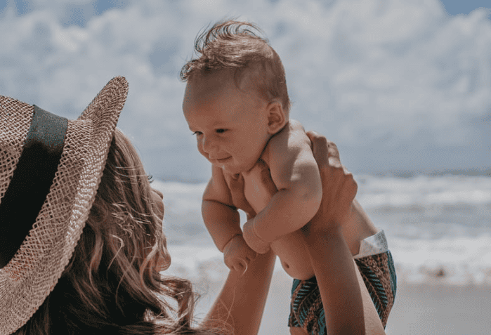 mom holding her baby in the air at the beach and experiencing summer as a new mom